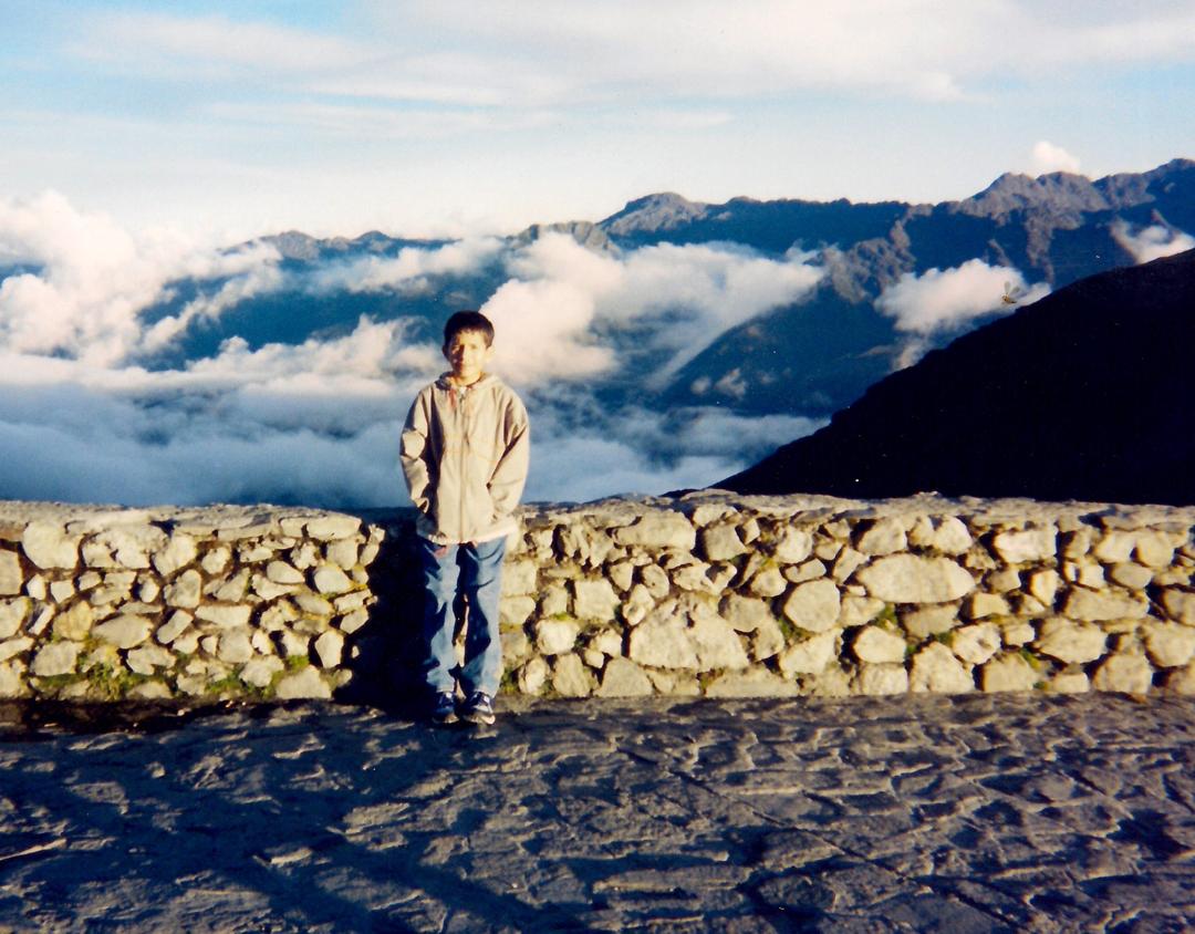 Carlos as a boy, standing on a stone terrace high in the mountains, a sea of clouds settled below the peaks behind him.